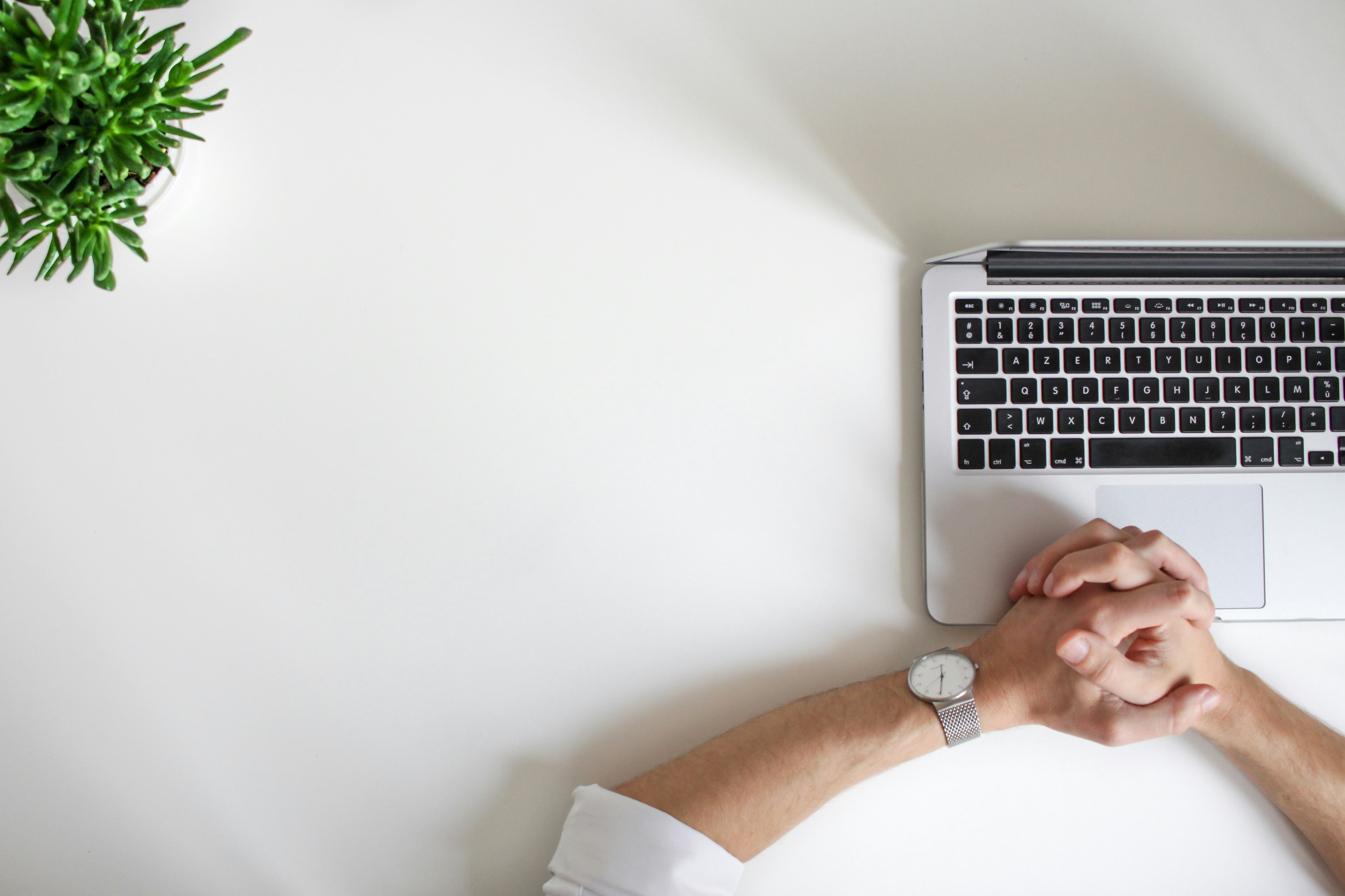 Two hands clasped over a laptop and a plant on the left corner